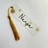 Marque-page transparent personnalisé avec le prénom « Neige », décoré de fleurs dorées et d’un pompon jaune.