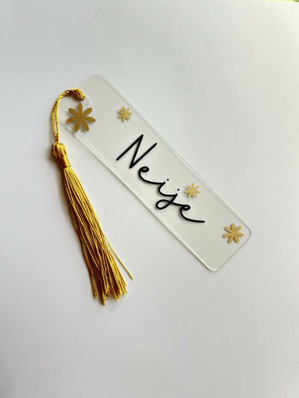 Marque-page personnalisé avec prénom et fleurs dorées – Idée cadeau originale Marque-page transparent personnalisé avec le prénom « Neige », décoré de fleurs dorées et d’un pompon jaune.